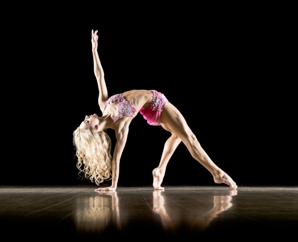 A female dancer in a pink costume performs a graceful backbend on a stage with one arm reaching upward, her blonde hair flowing, against a black background.