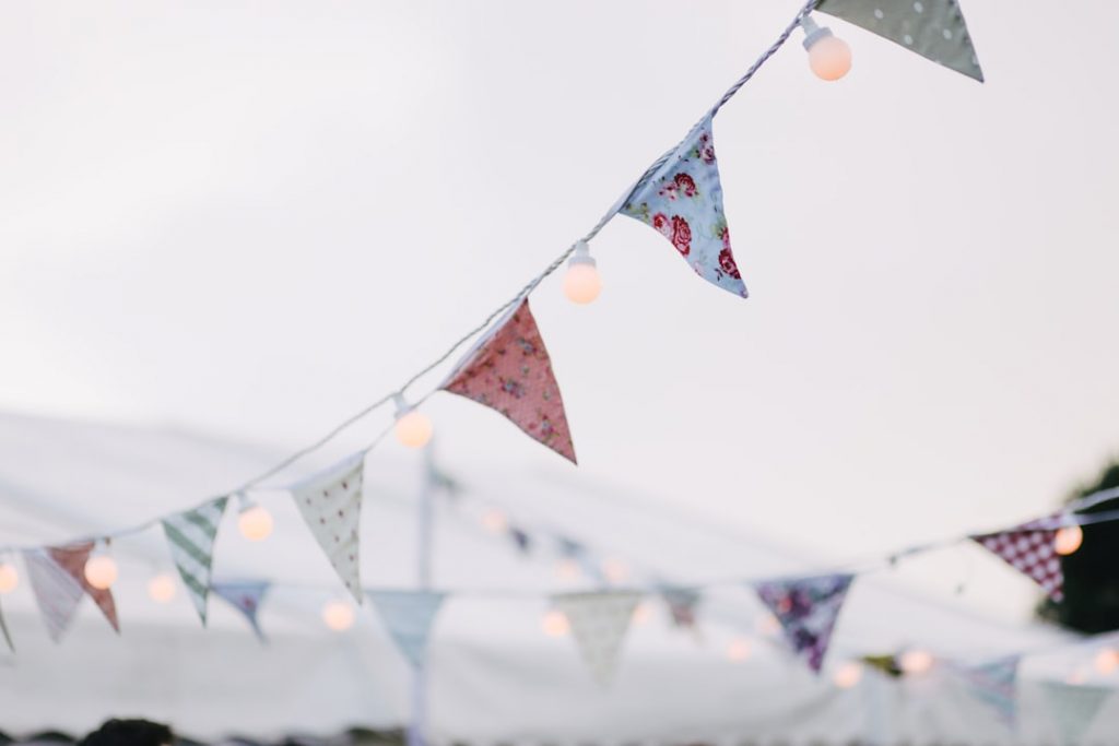 Colorful triangular bunting flags and string lights are hanging outdoors against a pale sky, creating a festive atmosphere. The background is soft and slightly out of focus.