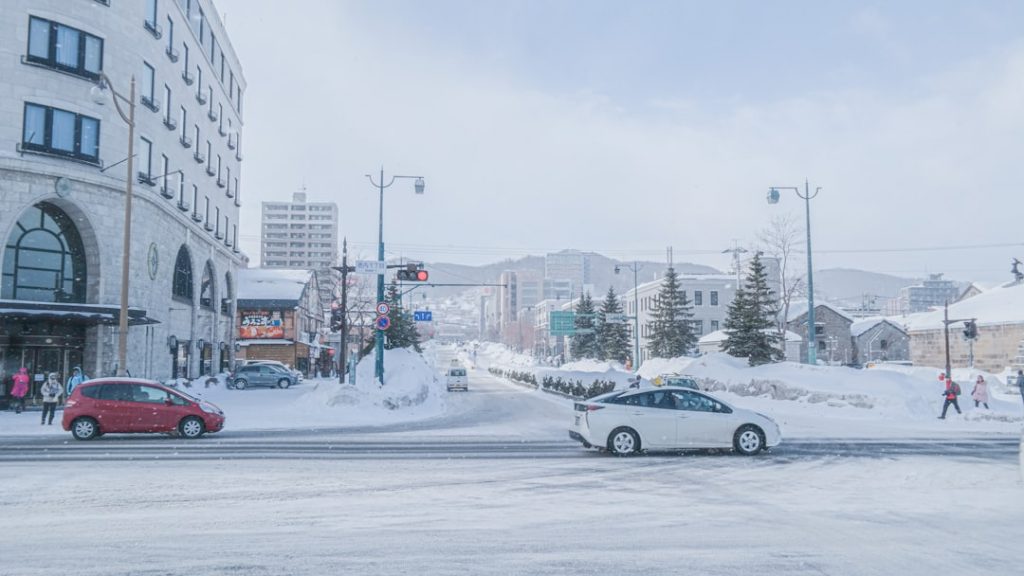 A snowy city street with cars driving and parked, buildings on both sides, and people walking. Snow covers the ground and trees, and distant hills are visible under a bright, slightly cloudy sky.