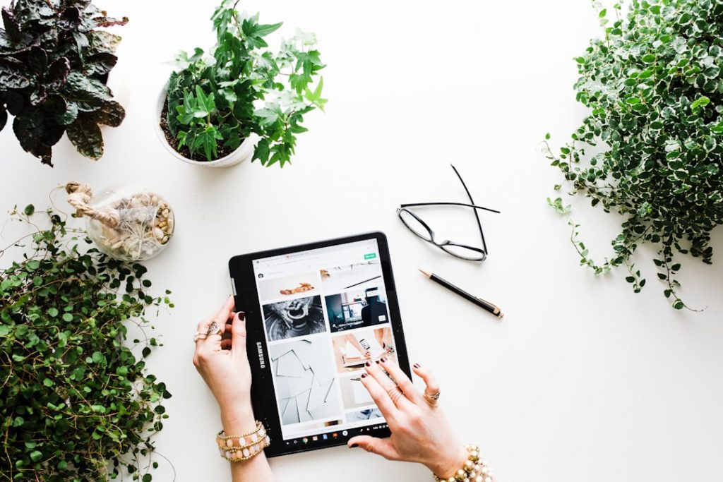 A person uses a tablet displaying images on a white desk surrounded by potted green plants, eyeglasses, and a pen. The person’s hands are visible, wearing bracelets and rings.