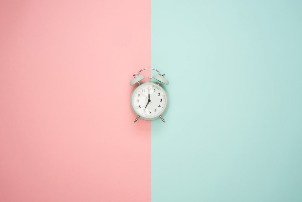 A small white alarm clock sits centered on a background split vertically into pink on the left and light blue on the right. The clock displays 10:10.