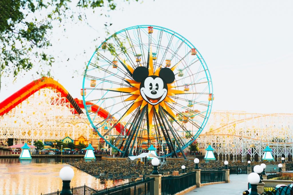 A large Ferris wheel with a smiling Mickey Mouse face at the center stands in a colorful amusement park, surrounded by water, lights, and roller coasters in the background.