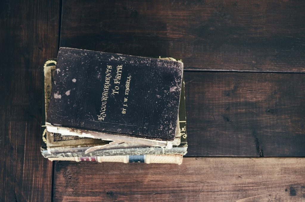 A stack of old, worn books sits on a dark wooden table. The top book, titled Encouragements to Faith by W. K. Tweedie, has a weathered black cover with visible scuff marks and frayed edges.