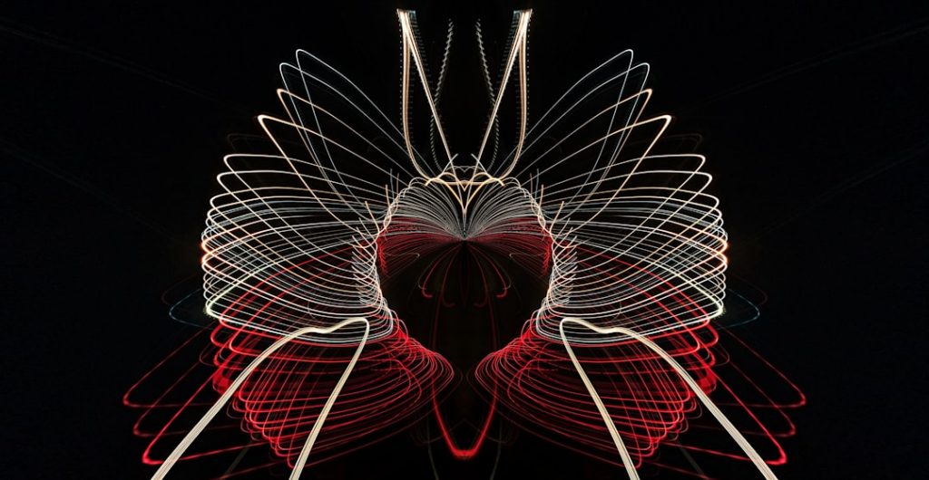 Abstract light painting with red, white, and yellow curved lines forming a symmetrical, wing-like pattern against a black background. The lines create a sense of movement and depth.