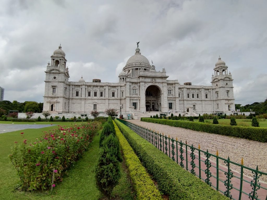 The image shows the grand white marble Victoria Memorial in Kolkata, India, with domes and towers, surrounded by manicured gardens and pathways under a cloudy sky.