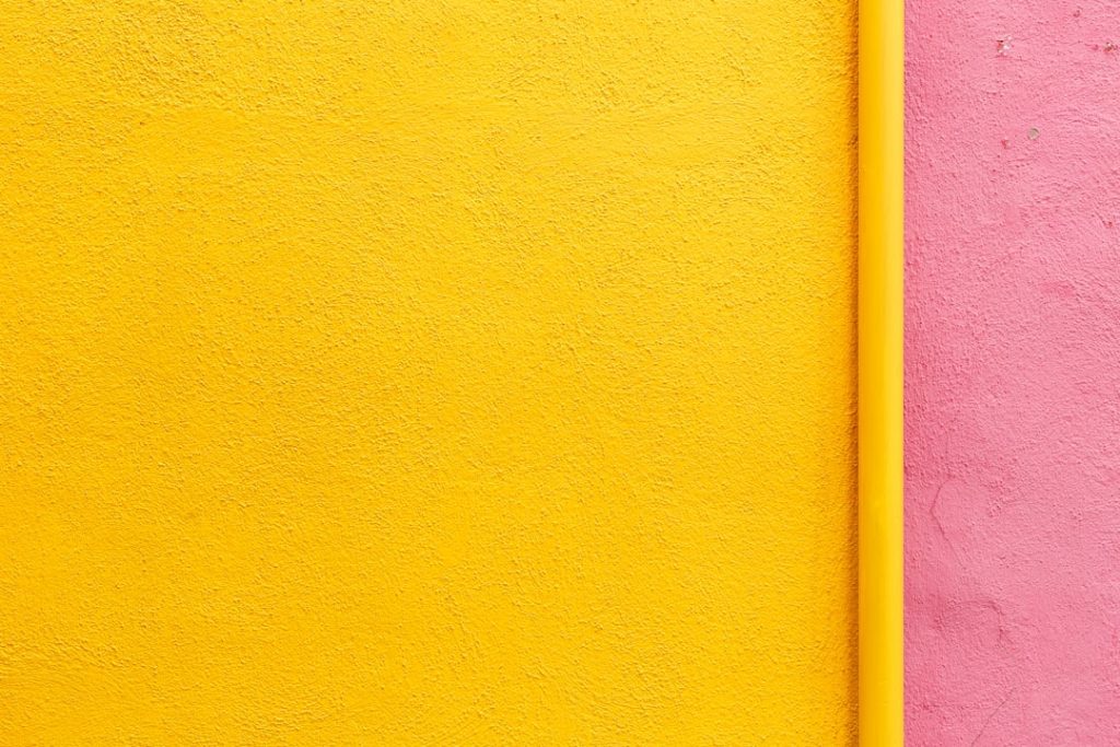 A textured wall divided vertically, with a bright yellow section on the left and a pink section on the right, separated by a yellow vertical pipe.