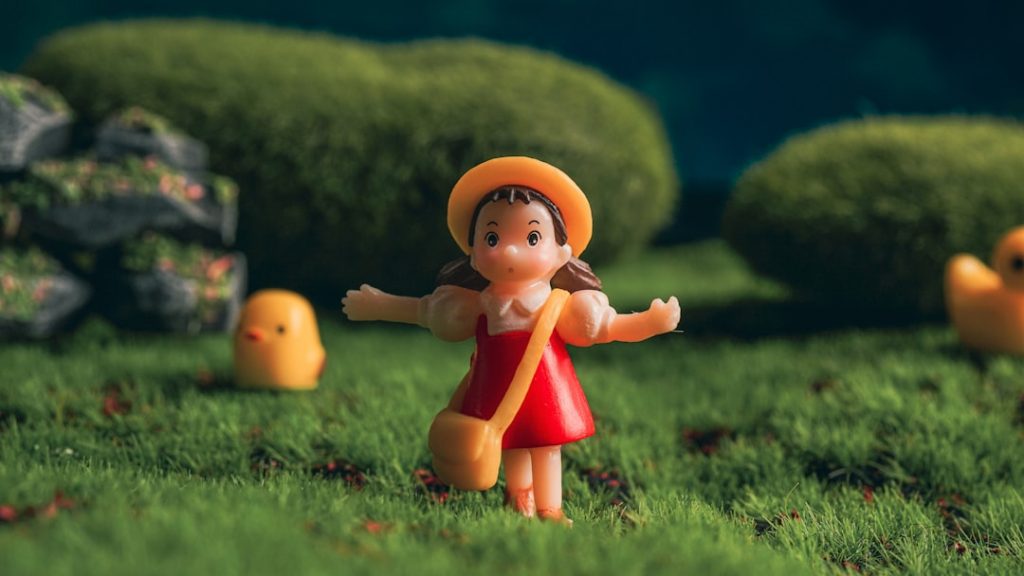 A small figurine of a girl with brown hair in pigtails, wearing a yellow hat, white shirt, red dress, and yellow bag, stands on green grass with toy ducks and bushes in the background.