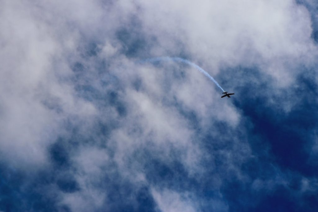 A small airplane flies through a cloudy blue sky, leaving a curved trail of white smoke behind it.