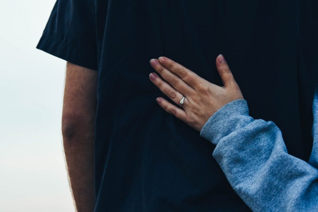 A person wearing a gray sleeve embraces another person in a black shirt from behind, with a hand resting on their back and an engagement ring visible on their finger.