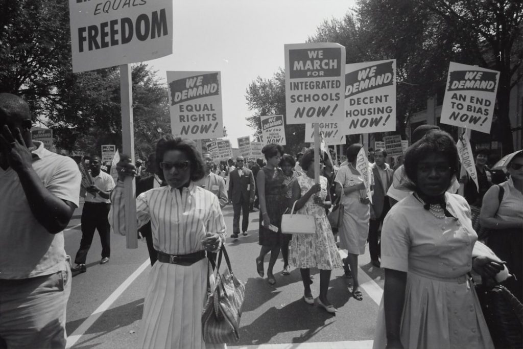 A group of civil rights activists march on a street holding signs demanding equal rights, integrated schools, decent housing, and an end to bias, during a 1960s protest.