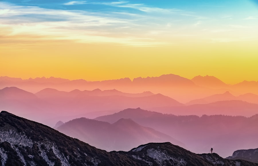 A vibrant sunset sky with orange, yellow, and blue hues above layers of misty mountains. A lone person stands on a rocky ridge in the foreground, gazing at the vast landscape.