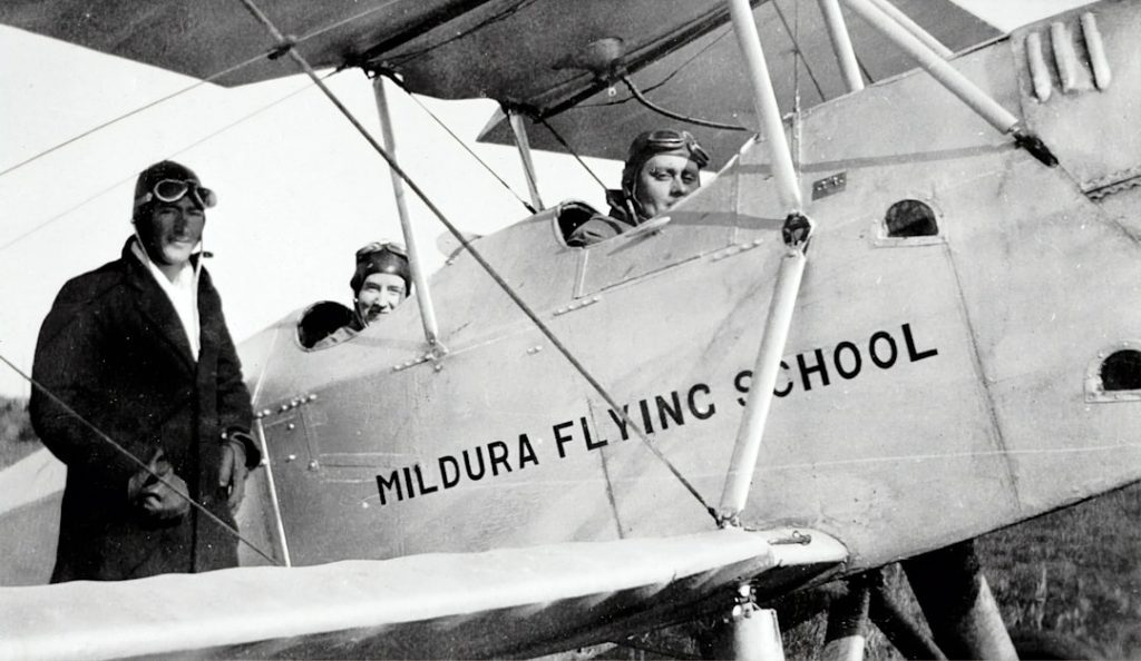 Three people in vintage flying gear pose with a biplane labeled Mildura Flying School. Two sit in the cockpit, while one stands beside the plane, all smiling at the camera. The image is in black and white.