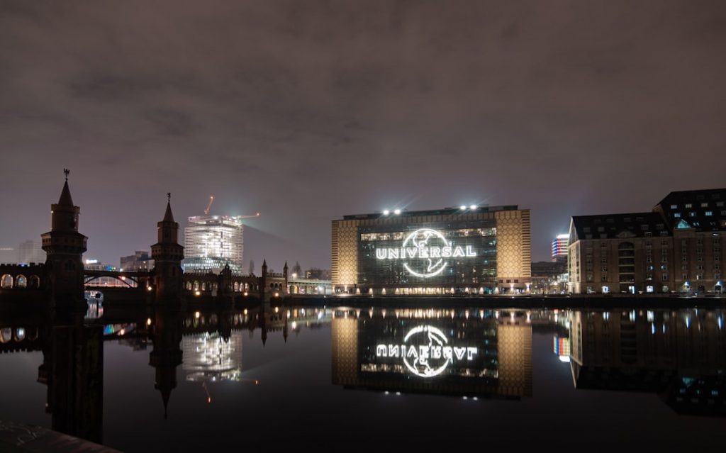 A nighttime cityscape featuring a river with buildings reflected on the water. The largest building displays the illuminated UNIVERSAL logo. A bridge with towers is visible on the left under a cloudy sky.