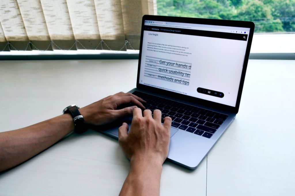 A person types on a laptop computer at a desk near a window with blinds. The laptop screen displays a webpage with text and navigation buttons.