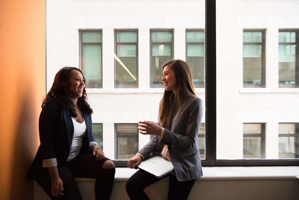 Two women in business attire sit by a large window, smiling and talking. One holds a laptop on her lap. They appear to be having a friendly conversation in a bright office setting.