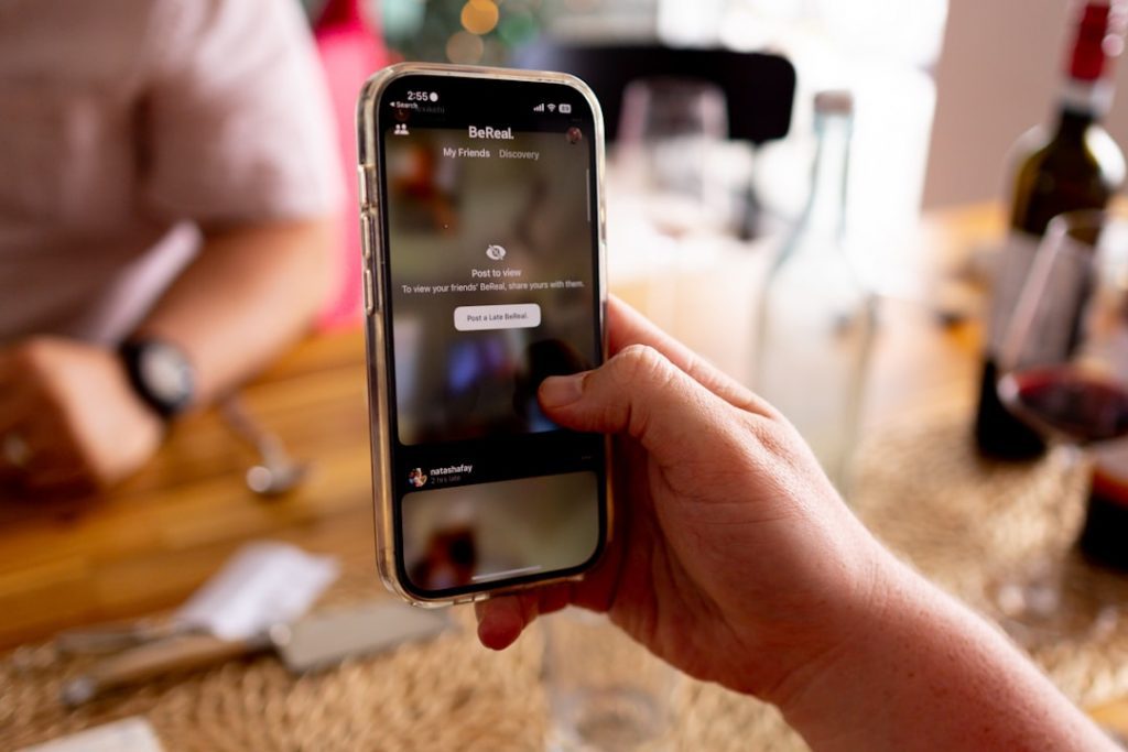 A hand holding a smartphone displaying the BeReal app, with a blurred dining table, plates, cutlery, and drinks in the background.
