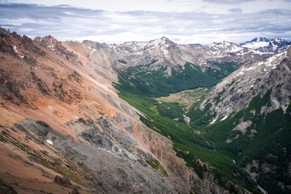 A panoramic view of a mountain valley with rugged, rocky slopes on both sides and a lush green forest stretching through the center under a cloudy sky.