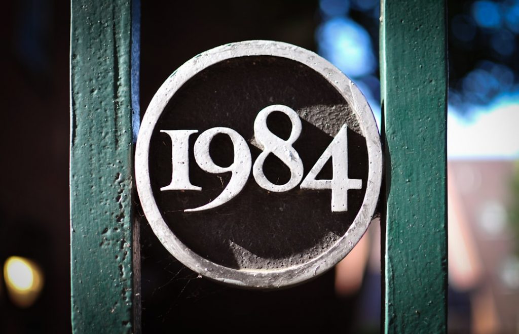 A close-up of a metal plaque with the number 1984 inside a circle, attached to a green metal fence. The background is slightly blurred.