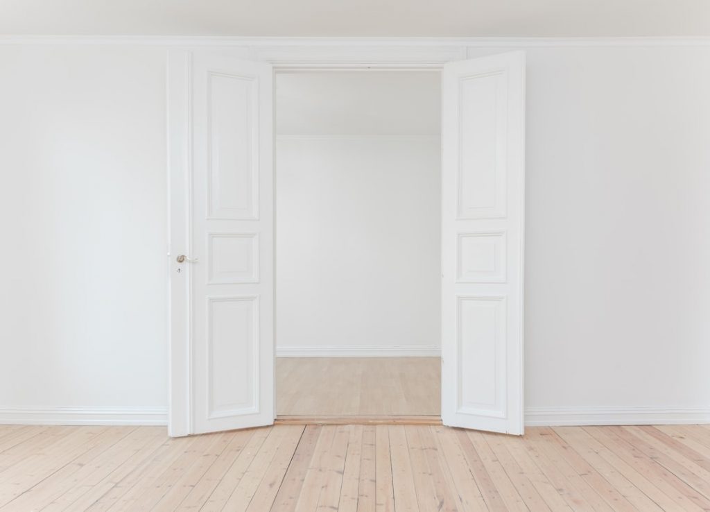 White double doors stand open in a minimalist room with white walls and light wood floors, leading into an adjoining empty space with similar decor. The area is bright and uncluttered.
