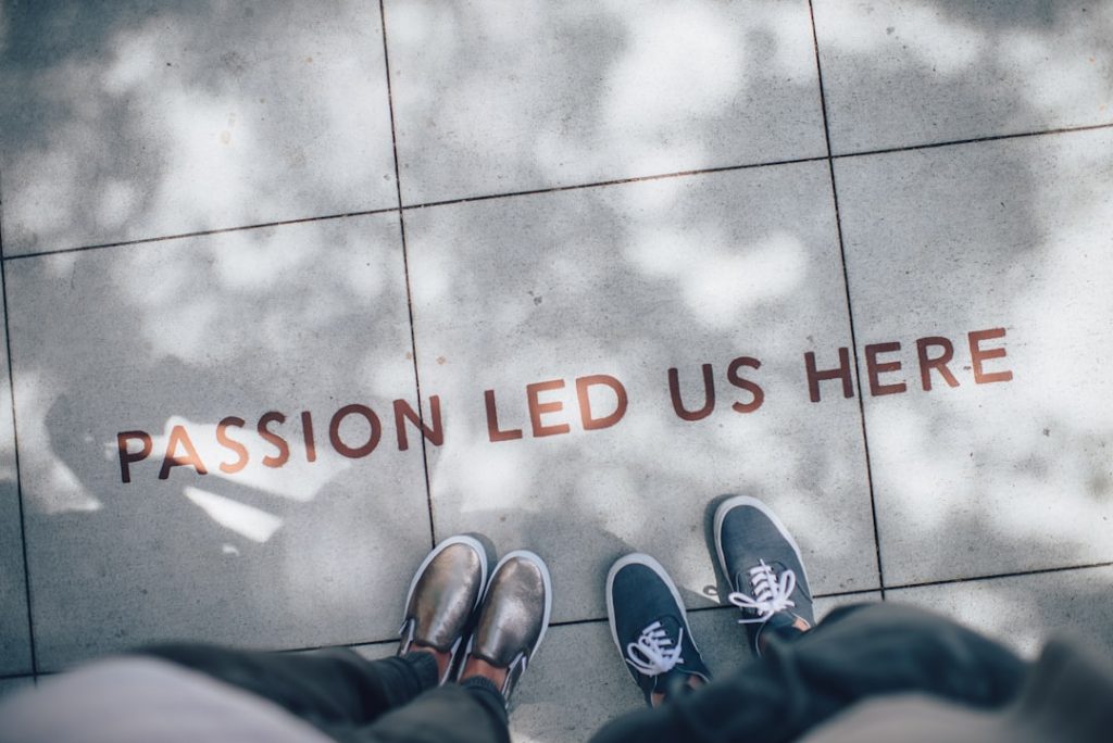 Two people standing on pavement next to each other, with the words PASSION LED US HERE written on the ground between them. Shadows of tree branches fall across the scene.