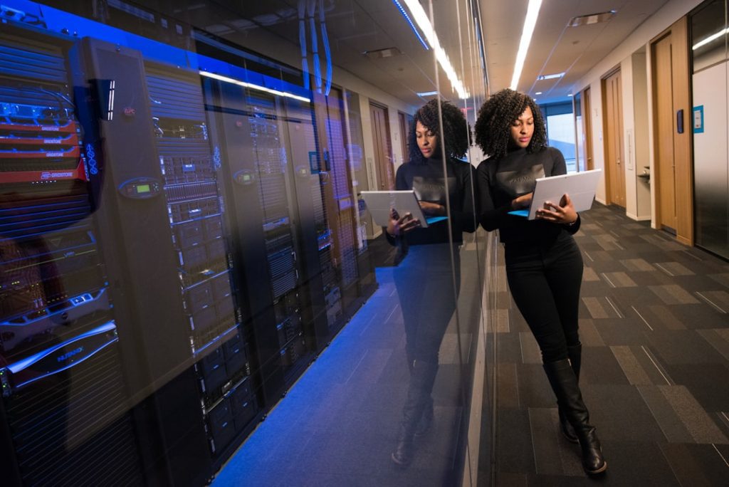 A woman stands in a modern server room, leaning against a glass wall with servers behind it, holding and working on a laptop. Her reflection is visible in the glass. The hallway is well-lit with overhead lights.