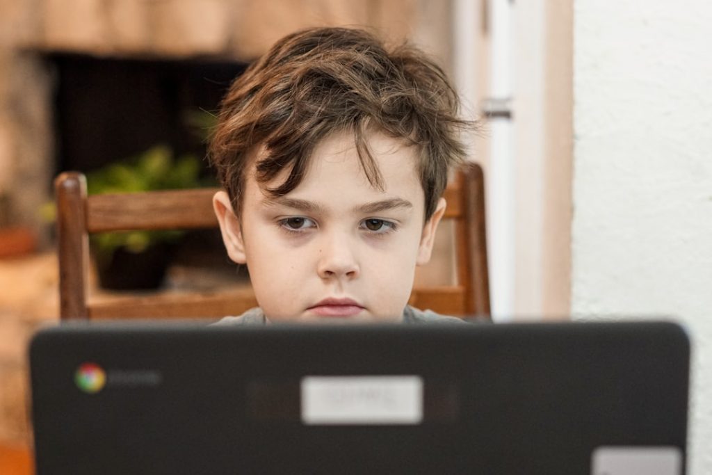 A young boy with brown hair sits at a table, focused on a laptop screen in front of him. The background is softly blurred, and the laptop has a Chrome logo on the back.