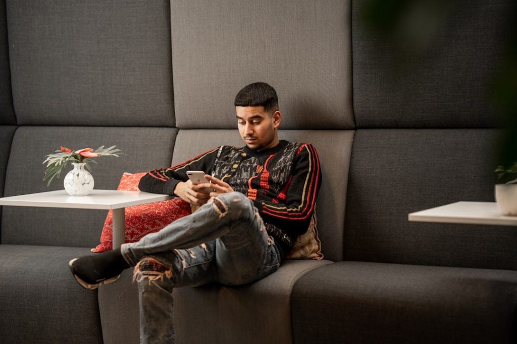 A man wearing a patterned jacket and ripped jeans sits on a gray cushioned bench, looking at his phone. There are small white tables with potted plants on either side of him.