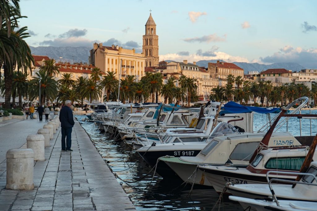 A row of boats docked along a waterfront promenade lined with palm trees, with people walking and historic buildings, including a tall bell tower, visible in the background under a partly cloudy sky.