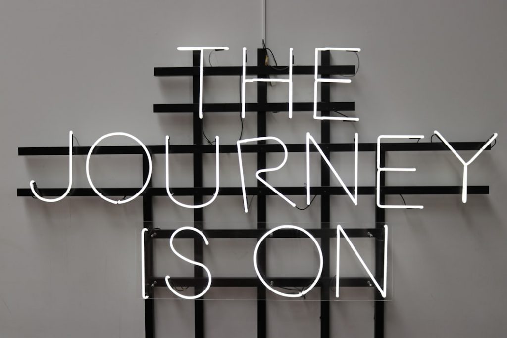 A white neon sign on a black metal grid reads, THE JOURNEY IS ON, set against a plain light gray wall.