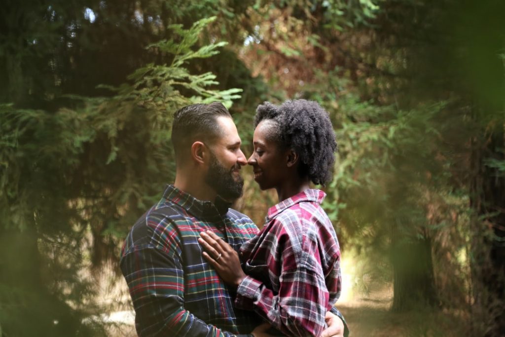 A couple wearing plaid shirts embraces and smiles at each other in a forest, surrounded by green trees and soft natural light.