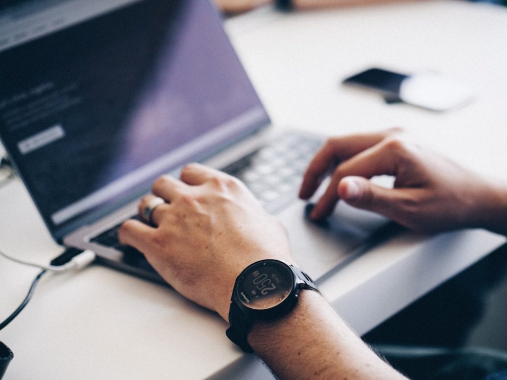 A person wearing a black smartwatch is typing on a laptop at a white desk, with a blurred smartphone in the background.