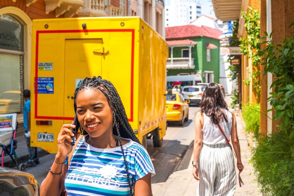 A woman with braided hair smiles while talking on her phone on a sunny street. A yellow truck and several cars are parked nearby, and another woman walks ahead in a striped dress. Buildings line both sides of the street.