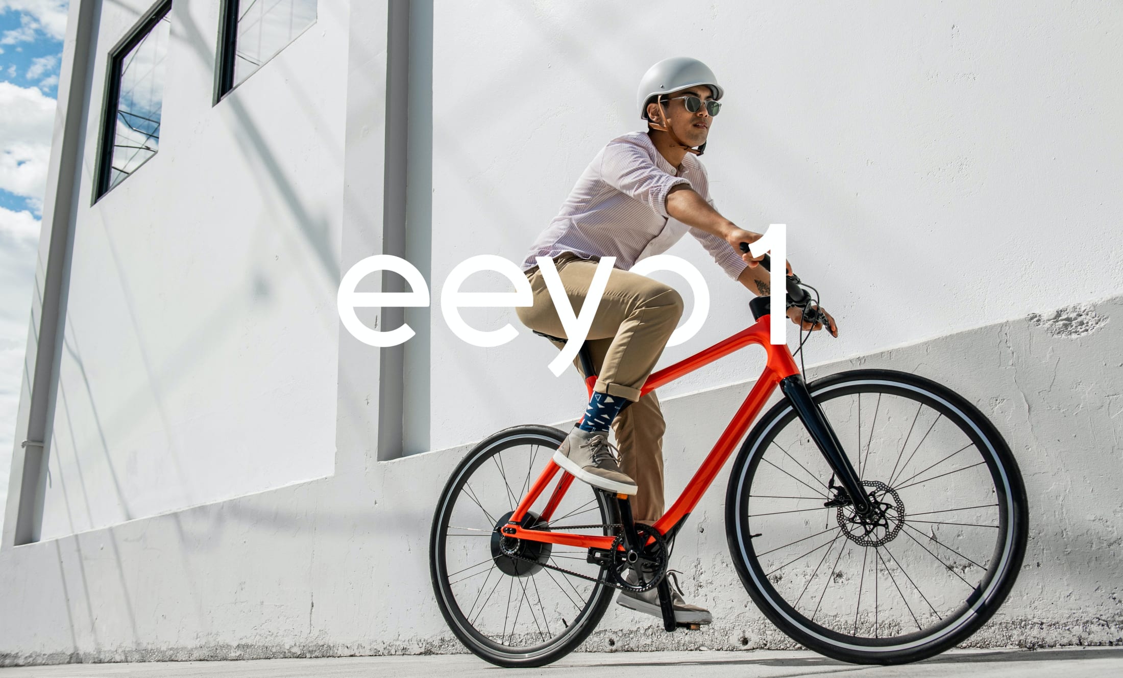 A person wearing a helmet and sunglasses rides a red bicycle on a sunny day, passing by a white building. The word “eeyo 1” is overlaid on the image.